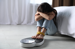 Person sitting on the floor looking distressed with feet on a scale and measuring tape — symbolizing emotional struggle with body image and eating disorder